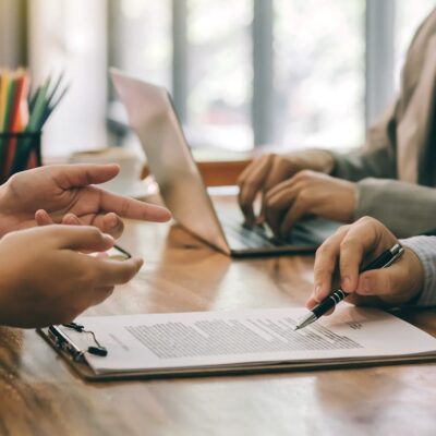 A Group Of People Sit At A Table With A Laptop And Clipboard, Reviewing Paperwork For A Business Loan.