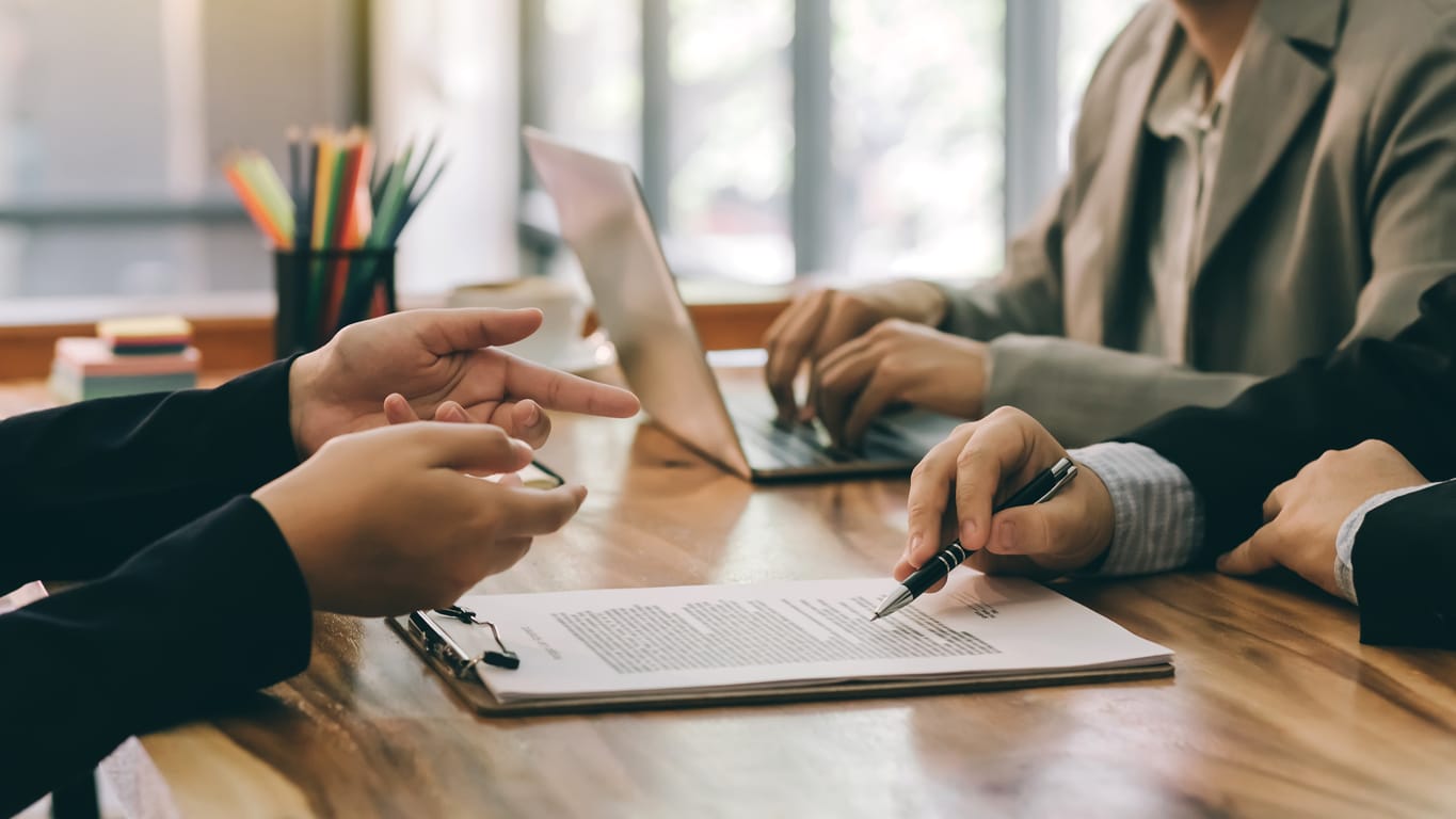 A group of people sit at a table with a laptop and clipboard, reviewing paperwork for a business loan.