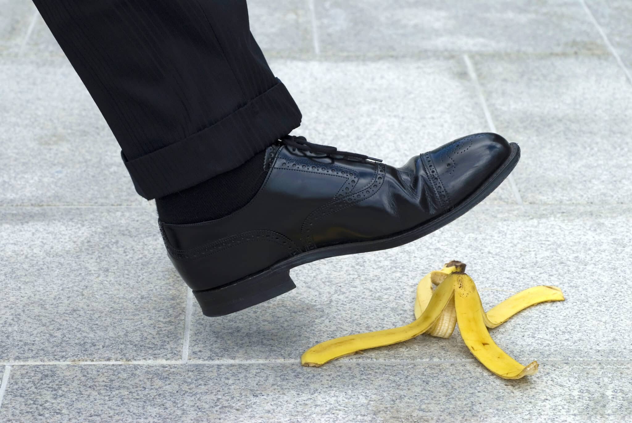 A man’s black brogue shoe poised to make the fateful mistake of stepping on a banana peel.